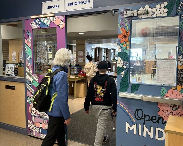 Student is using a long cane to walk into the library while the O&M Specialist is behind him. 