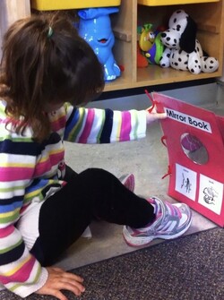 A student sits on the floor and holds a book about mirrors out at arms length. The open page has a mirror, print label, and pictures of ASL signs.