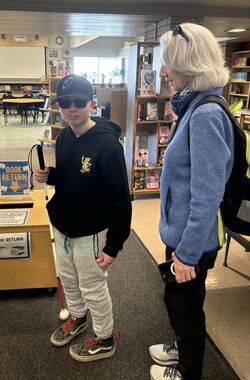 Student holds a long cane and looks at the camera while his O&amp;M specialist looks at the student.